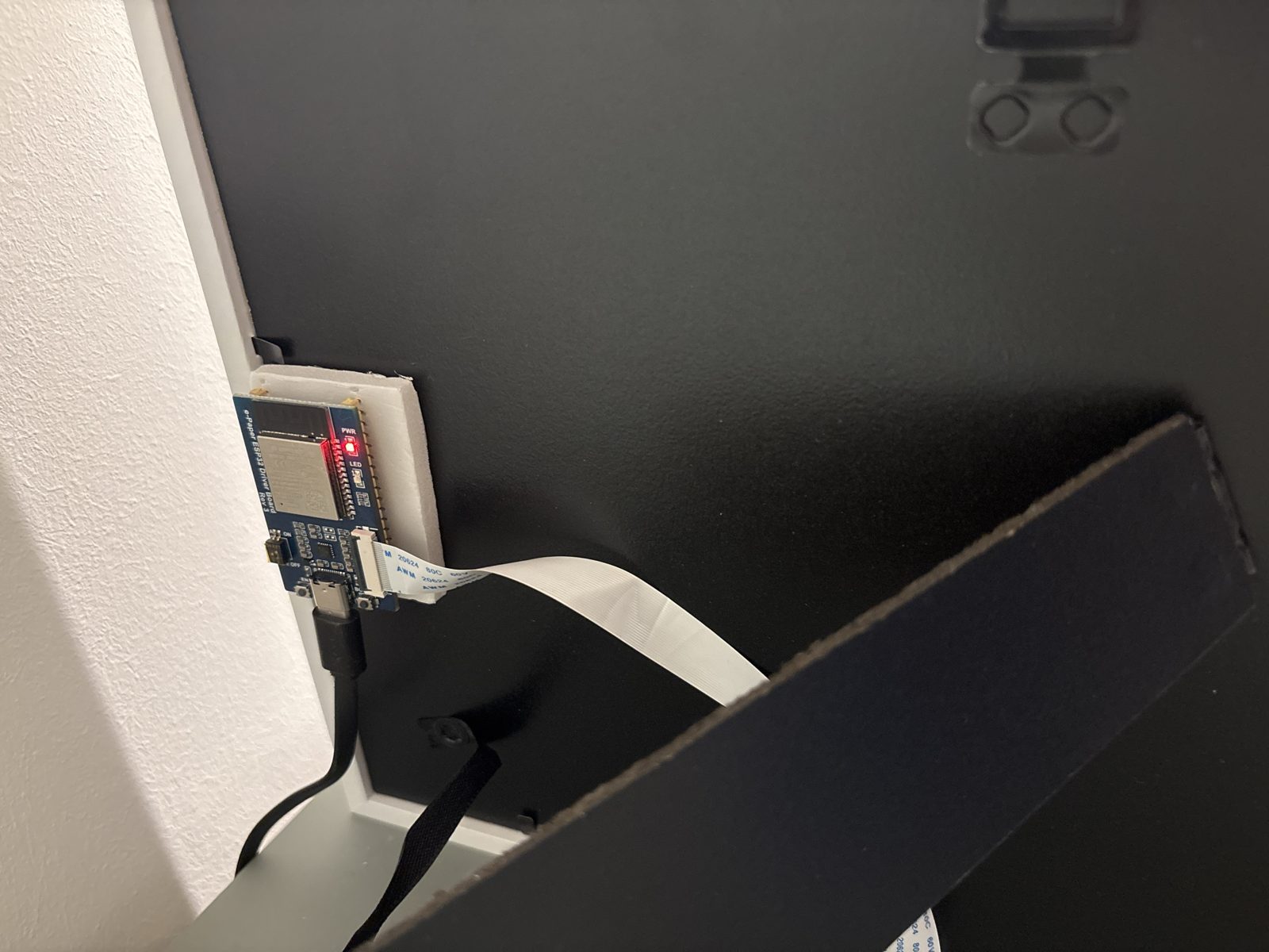 The back of the frame: the Waveshare ESP32 driver board, with its red power LED on, is stuck to a small foam pad. A white flat cable runs from the board to the screen, a USB-C cable hangs down to the power plug.